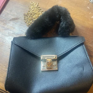 Svart handväska med pälsdetalj - Snygg svart handväska med en fluffig pälsdetalj på handtaget och en elegant guldkedja. Väskan har en stilren design med ett lås i guld framtill. Perfekt för att ge din outfit en lyxig touch. Helt oanvänd