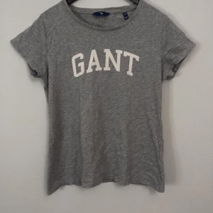 Grå t-shirt från GANT - Säljer en stilren grå t-shirt från GANT med vit text på framsidan. T-shirten är kortärmad och tillverkad i mjukt bomullsmaterial.