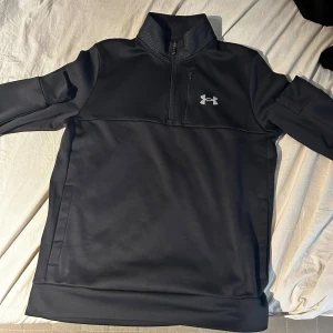 Svart tröja med dragkedja från Under Armour - Säljer en svart tröja från Under Armour med halv dragkedja och hög krage. Perfekt för träning eller vardagsbruk. Tröjan har en diskret logga på bröstet och är tillverkad i ett mjukt material.