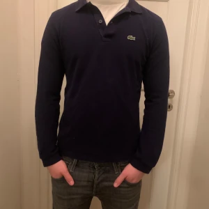 Mörkblå pikétröja från Lacoste - Säljer en stilren mörkblå pikétröja från Lacoste med klassisk krage och knappar. Tröjan har långa ärmar och den ikoniska krokodilloggan på bröstet. Perfekt för en avslappnad men ändå snygg look.