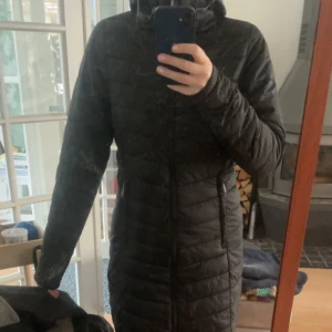 Svart lång dunjacka med huva - Säljer en svart lång dunjacka med huva. Jackan har en figurnära passform och är perfekt för kyliga dagar. Den har en dragkedja framtill och är quiltad för extra värme. Perfekt för vintern!