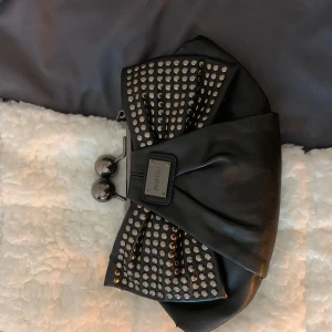 Svart kuvertväska från Fiorelli - Snygg svart kuvertväska från Fiorelli med glittriga detaljer och en stor rosett. Väskan har en metallisk stängning och en avtagbar axelrem. Perfekt för att ge en touch av glamour till din outfit.