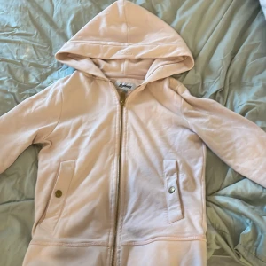 Beige huvtröja från Liseberg - Säljer en snygg beige huvtröja från Liseberg med dragkedja och praktiska fickor med knappar. Perfekt för en avslappnad stil. Tröjan har en broderad logga på huvan och är tillverkad i ett mjukt material.