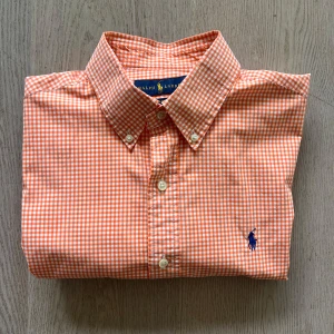 Orange rutig skjorta från Ralph Lauren - Snygg orange rutig skjorta från Ralph Lauren i slim fit. Skjortan har långa ärmar och klassisk krage med knappar. Den är tillverkad i bomull och har en broderad logga på bröstet. Perfekt för en stilren look!