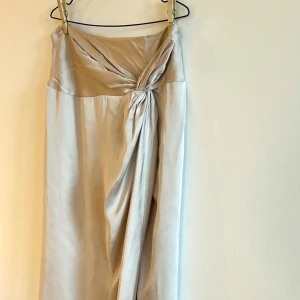 Helt oanvänd, silkig kjol från Stockh lm Studio - Elegant beige/ljust guldig kjol från Stockh lm Studio i ett silkigt material. Kjolen har en slits, en vacker drapering framtill och en knytning som ger en stilfull detalj. Helt oanvänd, lapparna kvar.