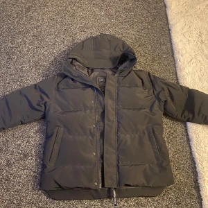 Svart pufferjacka med huva - Säljer en svart pufferjacka från canada goose Jackan har långa ärmar och en broderad logga på ärmen. Perfekt för kyliga dagar.