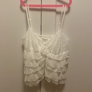 Vit spetsprydd linne med volanger - Söt vit linne med justerbara axelband och vackra spetsdetaljer. Toppen har flera lager av volanger som ger en romantisk känsla. Perfekt för varma sommardagar eller som ett lager under en kavaj. Den har även en öppning med två knappar överst som synligt på bilden.