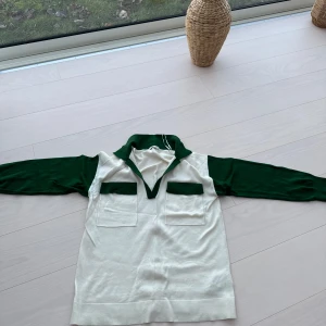 Vit och grön tröja med krage  - Snygg vit och grön tröja, krage med långa ärmar och två praktiska fickor framtill. Perfekt för en stilren och avslappnad look. Den har en ribbad nederkant och en unik design med gröna detaljer vid kragen och ärmarna.