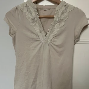 Beige topp med spetsdetaljer - Säljer en söt beige topp med spetsdetaljer runt den v-ringade halsen. Tight passform, saknar märke och storlek men skulle uppskatta att den är XS/S