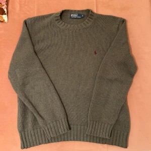 Olivgrön stickad tröja från Polo Ralph Lauren - Säljer en olivgrön stickad tröja från Polo Ralph Lauren. Tröjan har en klassisk rund hals och ett diskret broderat logotypmärke på bröstet. Tillverkad i 100% bomull, perfekt för kyligare dagar.