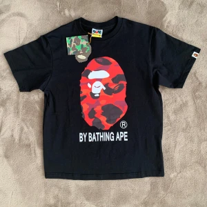 Svart BAPE T-shirt - Riktigt snygg svart t-shirt från Bape med det ikoniska röda och svarta kamouflagemönstret på framsidan. Den är helt ny och kommer med tags. Tröjan är i strl M men passar mer som en S. Vid frågor eller funderingar skicka ett pm, Mvh