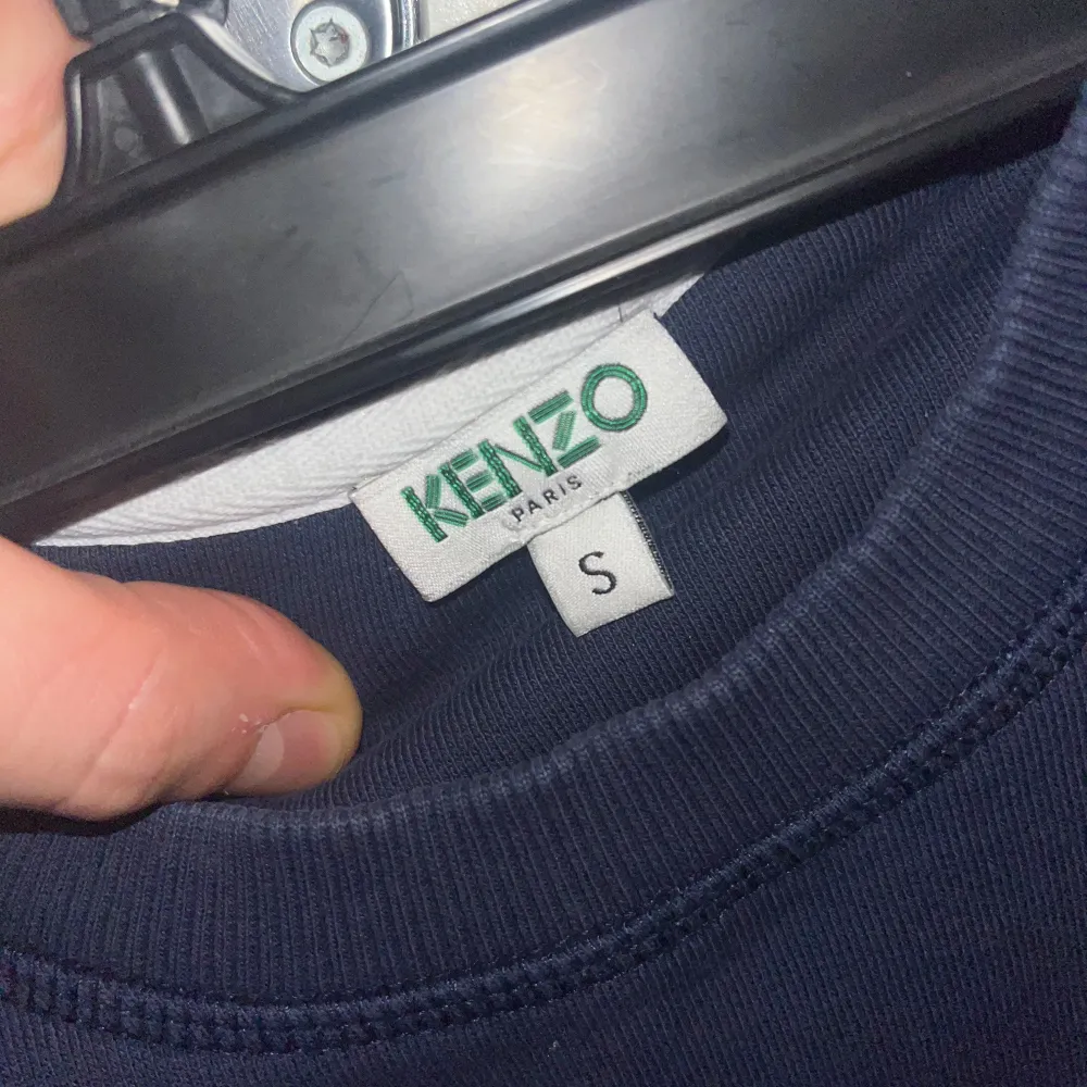 Säljer en mörkblå sweatshirt från Kenzo med ett stort tigerhuvud och texten 'Kenzo Paris' broderat på framsidan. Tröjan har långa ärmar och en rund halsringning. Perfekt för en cool och avslappnad stil. Tröjan är som ny och skulle sätta skicket som 9/10 med tanke på vardaglig användning😊. Neuletakit & Villapaidat.