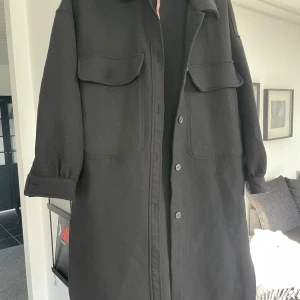 Svart kappa med knappar - Säljer en stilren svart kappa i mycket bra skick. Den har långa ärmar och knappar framtill. Perfekt för höst och vår, med stora fickor för extra stil och funktionalitet. Passar både till vardags och lite finare tillfällen.