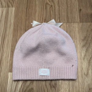 Rosa stickad mössa från UGG - Söt rosa stickad mössa från UGG med en fin rosett på toppen. Perfekt för att hålla värmen under kyliga dagar. Den har en ribbad kant för extra komfort och stil.