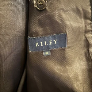 Svart rock från Riley - Säljer en svart rock från Riley i storlek 52. Rocken har en klassisk design med knappar framtill och är perfekt för både formella och semi-formella tillfällen. Materialet känns lyxigt och passar bra för höst och vinter. Perfekt för att ge en stilren look!