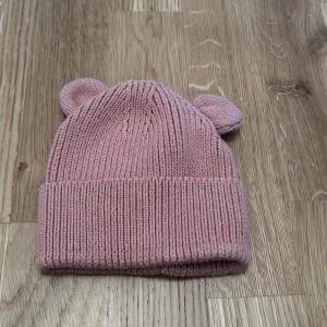Rosa stickad mössa med öron från H&M - Söt rosa stickad mössa från H&M med små öron på toppen. Perfekt för att hålla de små varma och stiliga under kyliga dagar.