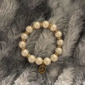 Elegant armband med vita pärlor och guldfärgade detaljer. Armbandet har en charmig peace-berlock i guldton som ger en bohemisk touch. Perfekt för att lyfta vilken outfit som helst.
