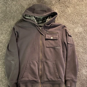 Fur/fluffig hoodie - Mysig svart hoodie från s.Oliver med dragkedja och fodrad med mjuk pälsimitation. Den har en praktisk huva och flera fickor med knappdetaljer. Perfekt för kyliga dagar. Storlek M