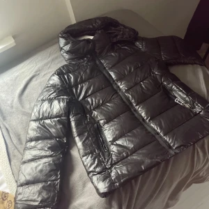Svart pufferjacka med huva - Säljer en snygg svart pufferjacka i nytt skick. Jackan har en glansig yta och är perfekt för kalla dagar. Den har en praktisk huva och dragkedja framtill. Perfekt för vintern och hösten! den är i Xl men passar mer som en L