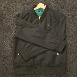 Mörk blå bomberjacka från GANT - Säljer en snygg blå bomberjacka från GANT i bra skick. Jackan har en dragkedja framtill och två fickor med knappar. Perfekt för höst och vår, med en stilren design som passar till det mesta. Inuti är den fodrad med en turkos färg som ger en cool kontrast.