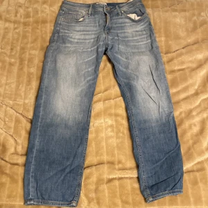 Blå jeans från Jack & Jones - Säljer ett par klassiska blå jeans från Jack & Jones i modellen Regular/Clark. De har en normal passform med dragkedja och knapp framtill. Perfekta för vardagsbruk och passar till det mesta. Jeansen är i bra skick och redo för en ny ägare!
