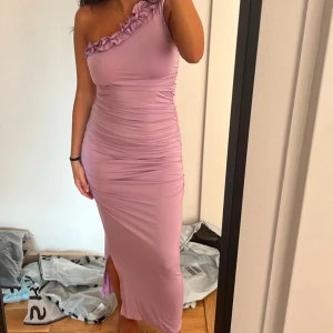 Ljuslila oneshoulder klänning - Elegant ljuslila oneshoulder klänning med volangdetaljer vid axeln. Klänningen har en smickrande passform med slits på sidan, perfekt för speciella tillfällen.