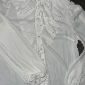 vit blus med volanger - säljer en jättesöt och elegant blus med volanger från Shein. blusen är perfekt både för vardag och fest. mycket bra skick, använd ett fåtal ggr. väldigt genomskinlig dock men funkar fint med ett linne under då den är väldigt lätt och skön. (OBS!! liten i storleken, passar S) 
