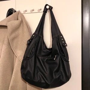 Svart axelväska med nitar - Snygg svart axelväska i skinn med coola nitar och dragkedja. Väskan e köpt secondhand och har därför några defekter. Insidan av väskan är lite trasig och det finns sågspån på insidan (😭) utöver det funkar väskan prima. Super trendig dessutom. Liknar vid den stilen av väska på sista bilden😍