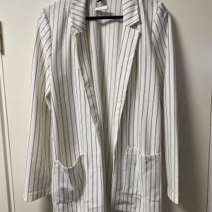 Vit randig kavaj - Säljer en stilren vit kavaj med svarta ränder. Den har en klassisk design med långa ärmar och två fickor framtill. Perfekt för både vardag och festliga tillfällen. Passar bra till både jeans och mer formella byxor.