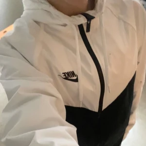 Vit och svart vindjacka från Nike - Säljer en snygg vit och svart vindjacka från Nike i storlek S. Jackan har en dragkedja framtill och är perfekt för vår och höst. Den är i mycket bra skick och har en klassisk Nike-logga på bröstet. Perfekt för träning eller vardagsbruk!