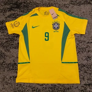 Gul Brasilien fotbollströja - Säljer en gul Brasilien fotbollströja från Nike med gröna detaljer och nummer 9 på framsidan. På baksidan står det 'Ronaldo' med samma nummer. Tröjan är kortärmad och i nytt skick, perfekt för samlare eller fans av det brasilianska landslaget. Passar perfekt för matchdagar eller som en cool vardagströja!