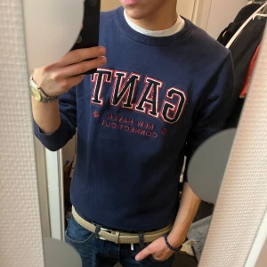 Mörkblå sweatshirt från GANT - Säljer en snygg mörkblå sweatshirt från GANT med broderad logga i rött och vitt. Tröjan är i bra skick och perfekt för en casual look. Den har en klassisk rund hals och är superbekväm. Passar perfekt till jeans för en avslappnad stil.