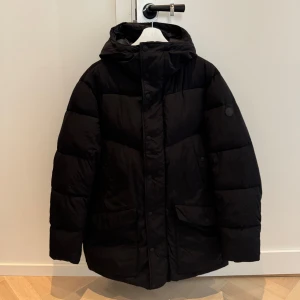 Svart pufferjacka från Save the Duck - Säljer en svart pufferjacka från Save the Duck. Jackan är djurvänlig och har en quiltad design med lång ärm och huva. Perfekt för kalla dagar. Endast använd ett fåtal gånger.