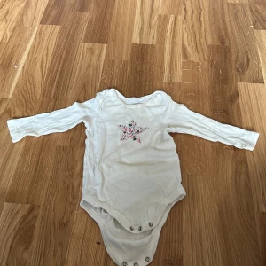 Vit långärmad body från Lupilu - Söt vit långärmad body från Lupilu med en stjärndetalj i blommigt mönster på framsidan. Perfekt för de små under vår och sommar. Praktiska knappar i grenen för enkel på- och avklädning.