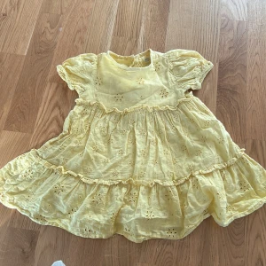 Gul klänning från Fred & Flo - Söt gul klänning från Fred & Flo i bomull med broderade blommor och volanger. Klänningen har korta ärmar och knappar i ryggen för enkel påklädning. Perfekt för varma sommardagar!