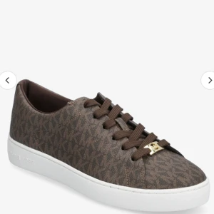 Bruna sneakers från Michael Kors - Snygga bruna sneakers från Michael Kors med ett diskret mönster och vita sulor. Skorna har snörning och en elegant design som passar perfekt till vardags.