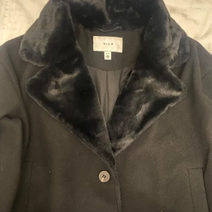 Svart kappa från VILA - Elegant svart kappa från VILA med mjuk pälsimitation på kragen. Perfekt för kyliga dagar med sin stilrena design och knäppning framtill. Ett måste för garderoben!