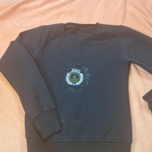 Svart tröja med Judo-märke - Snygg svart tröja med ett coolt Judo-märke på bröstet, omgiven av glittrande detaljer. Perfekt för dig som älskar kampsport och vill ha en unik stil. Tröjan har långa ärmar och en bekväm passform.
