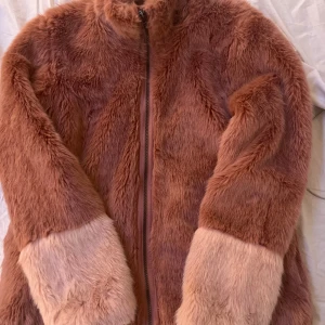Rosa pälsjacka med dragkedja - Mysig rosa pälsjacka med dragkedja och långa ärmar. Jackan har en fluffig textur och är perfekt för kyliga dagar. Den har en ljusare nyans vid ärmsluten som ger en fin kontrast. Den är i storlek 170 men skulle säga att den är en stor s/liten m 