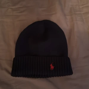 Mörk blå mössa från Ralph Lauren - Snygg mörk blå mössa från Ralph Lauren med röd broderad logotyp. Den är ribbad och tillverkad i mjukt material, perfekt för kyliga dagar. Passar de flesta huvudstorlekar.