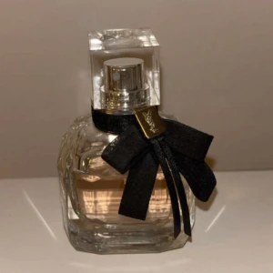 Parfym med svart rosett - Säljer en elegant parfymflaska med en svart rosett och gulddetaljer. Flaskan är fyrkantig och genomskinlig, vilket visar den ljusrosa vätskan inuti. Perfekt för den som vill ha en stilren och lyxig doft på hyllan.