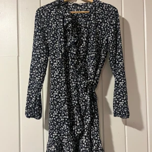 Blommig klänning med volangdetaljer - Säljer en blommig klänning i mörkblått med vita blommor. Den har långa ärmar och volangdetaljer framtill som ger en romantisk touch. Perfekt för vår och sommar! Klänningen har en knytning i midjan för en smickrande passform.