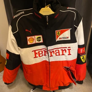 Ferrari jacka (OBS Lite trasig) - Snygg Ferrari jacka i svart, rött och vitt med flera broderade loggor som Shell och UPS. Jackan har dragkedja och är långärmad med en cool racingstil. Perfekt för bilentusiaster!