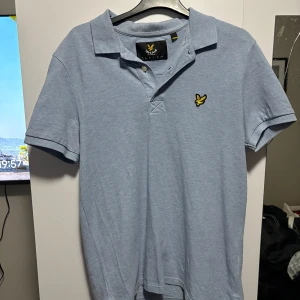 Ljusblå pikétröja från Lyle & Scott - Säljer en stilren ljusblå pikétröja från Lyle & Scott med klassisk krage och knappar. Tröjan har en broderad gul logotyp på bröstet. Perfekt för en avslappnad look.