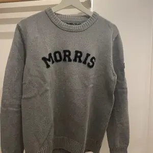 Säljer en snygg grå stickad tröja från Morris med texten 'MORRIS' i svart på bröstet. Tröjan har långa ärmar och ribbade muddar vid ärmslut och nederkant. Perfekt för kyliga dagar och en stilren look. 🇬🇧