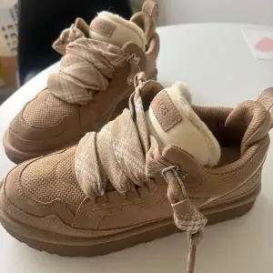 Säljer ett par snygga beige sneakers från UGG med fluffigt foder och breda snören. Skorna har en robust sula och är perfekta för kyligare dagar. De är både stiliga och bekväma. Färg Sand