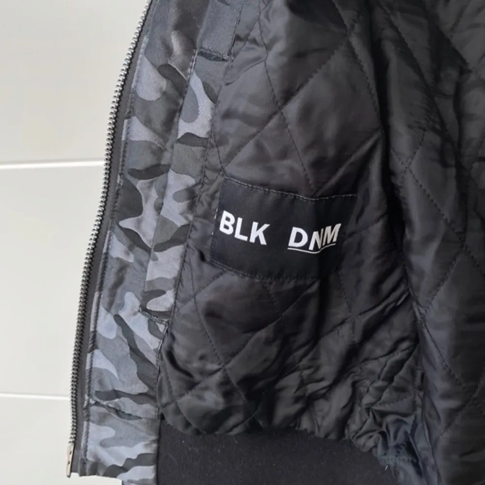 Snygg kort bomberjacka i grått camouflage från BLK DNM. Jackan har en dragkedja framtill och ribbade muddar vid ärmslut och nederkant. Perfekt för en cool och avslappnad stil.. Takit.