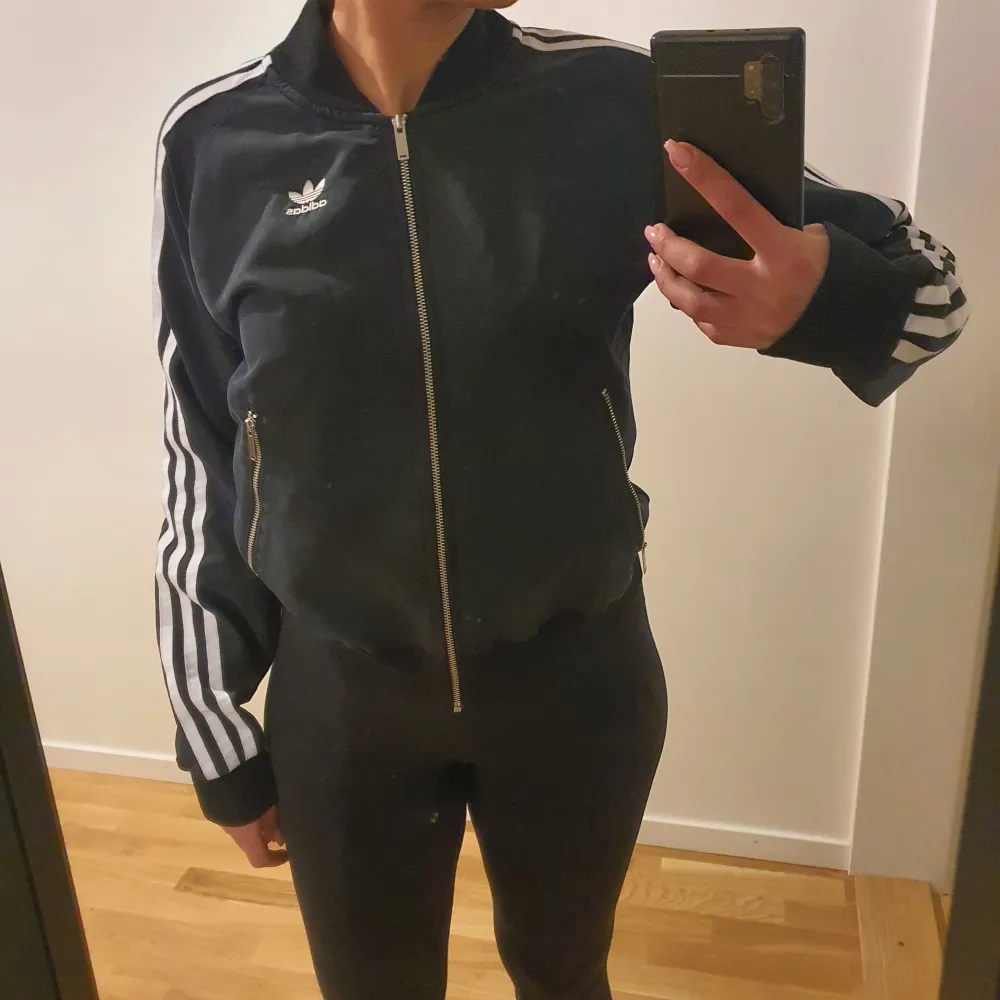 Snygg svart koftjacka från Adidas med klassiska vita ränder längs ärmarna. Jackan har dragkedja framtill och två fickor med dragkedjor. Perfekt för en sportig look.. Tröjor & Koftor.