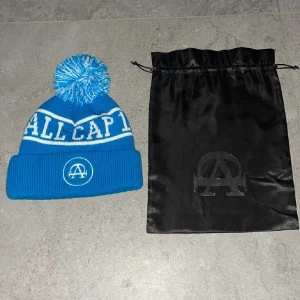 Allcap one mössa - Snygg blå mössa med stor tofs och texten 'ALL CAP' i vitt. Mössan har en ribbad kant och ett tryckt emblem framtill. Perfekt för kalla dagar!     Pris kan diskuteras     Ny pris 300kr påse får man med