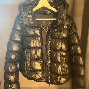 Svart pufferjacka med huva - Säljer min svarta glansiga superdry jacka då den inte kommer komma till användning mer. Inte speciellt använd, inga tecken på skada. Varm och skön i höst o vinter!!